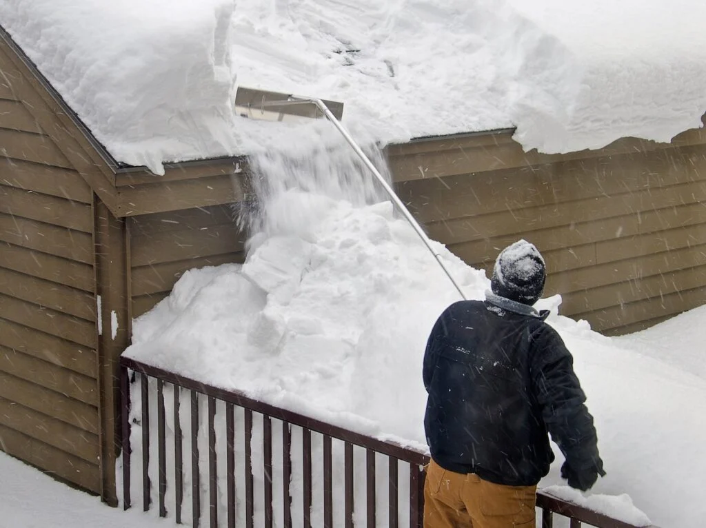homeowner using a roof rake from the ground to clear snow off the eaves. 