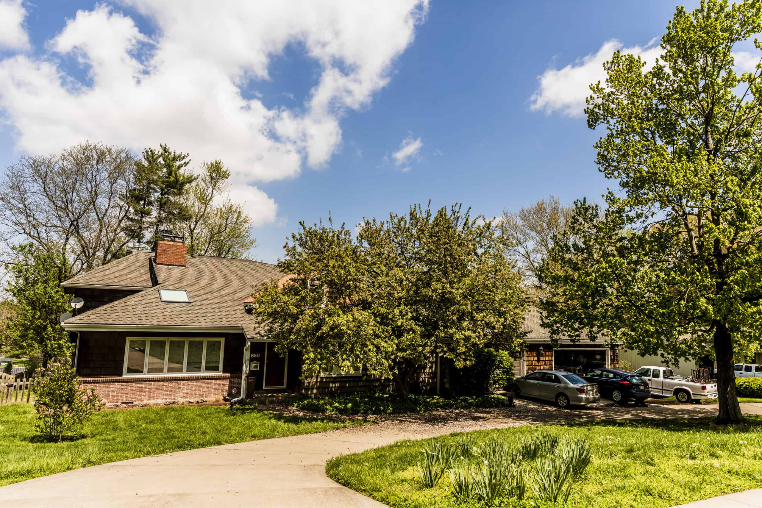 Gable roof with shingle roofing, trees, cars, and a clear blue sky in a residential neighborhood.