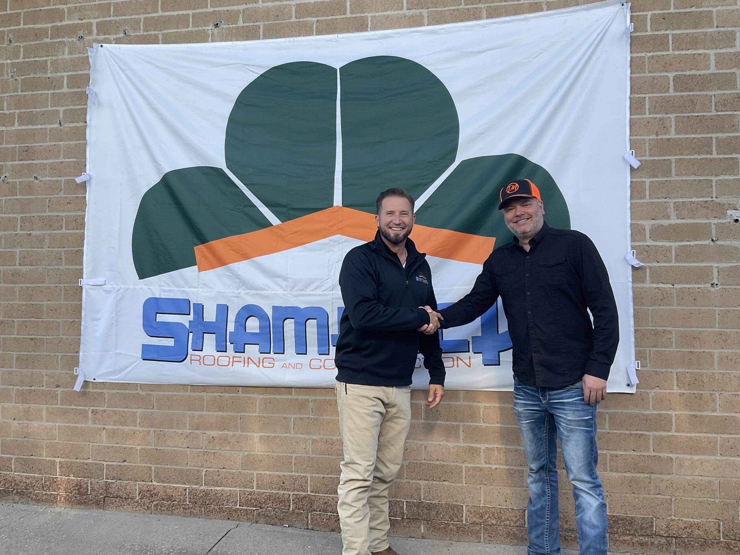 Shamrock Roofing and Construction team members shaking hands in front of company banner on brick wall.